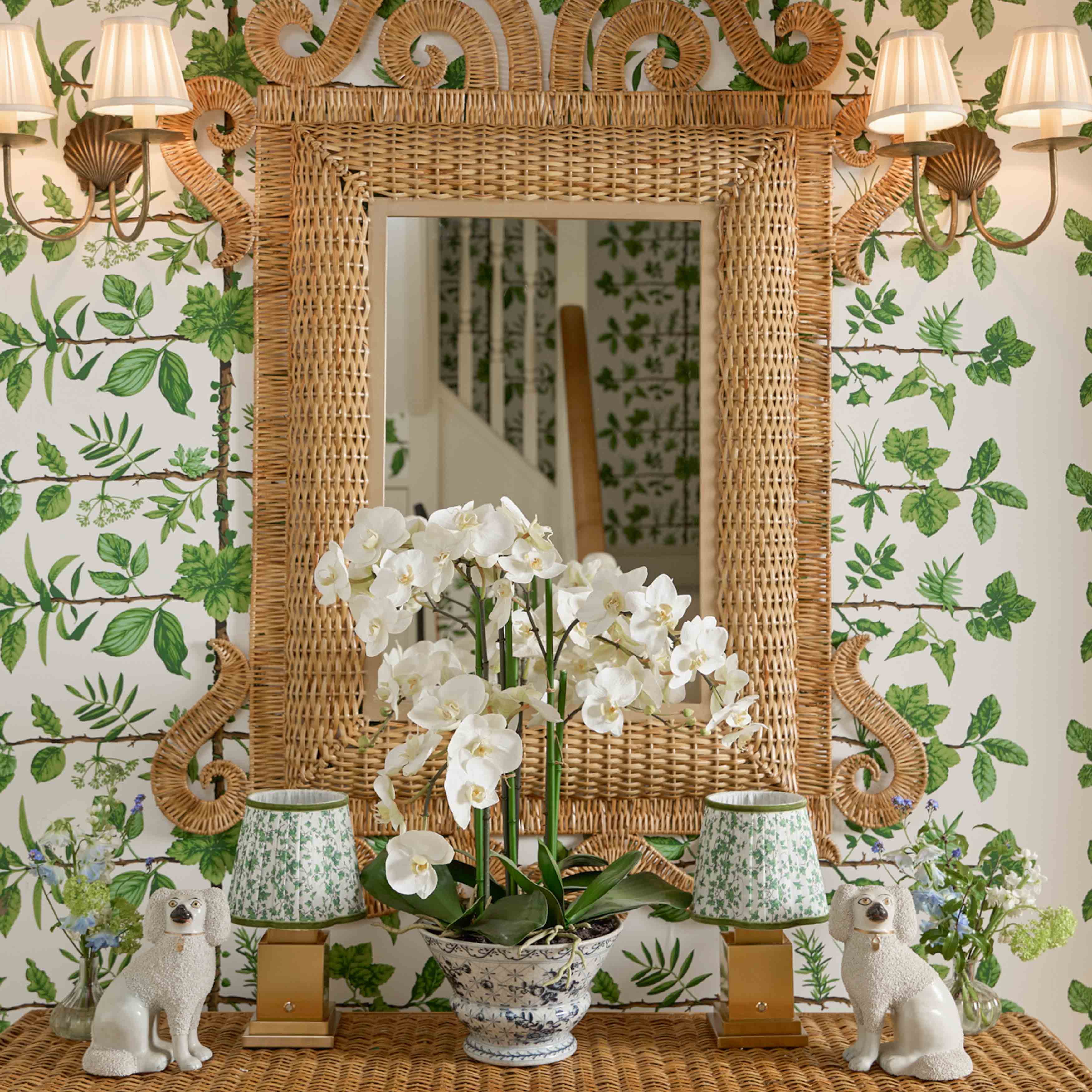 Decorative mirror with wicker frame, white orchids, and small lamps against a leaf-patterned wallpaper.