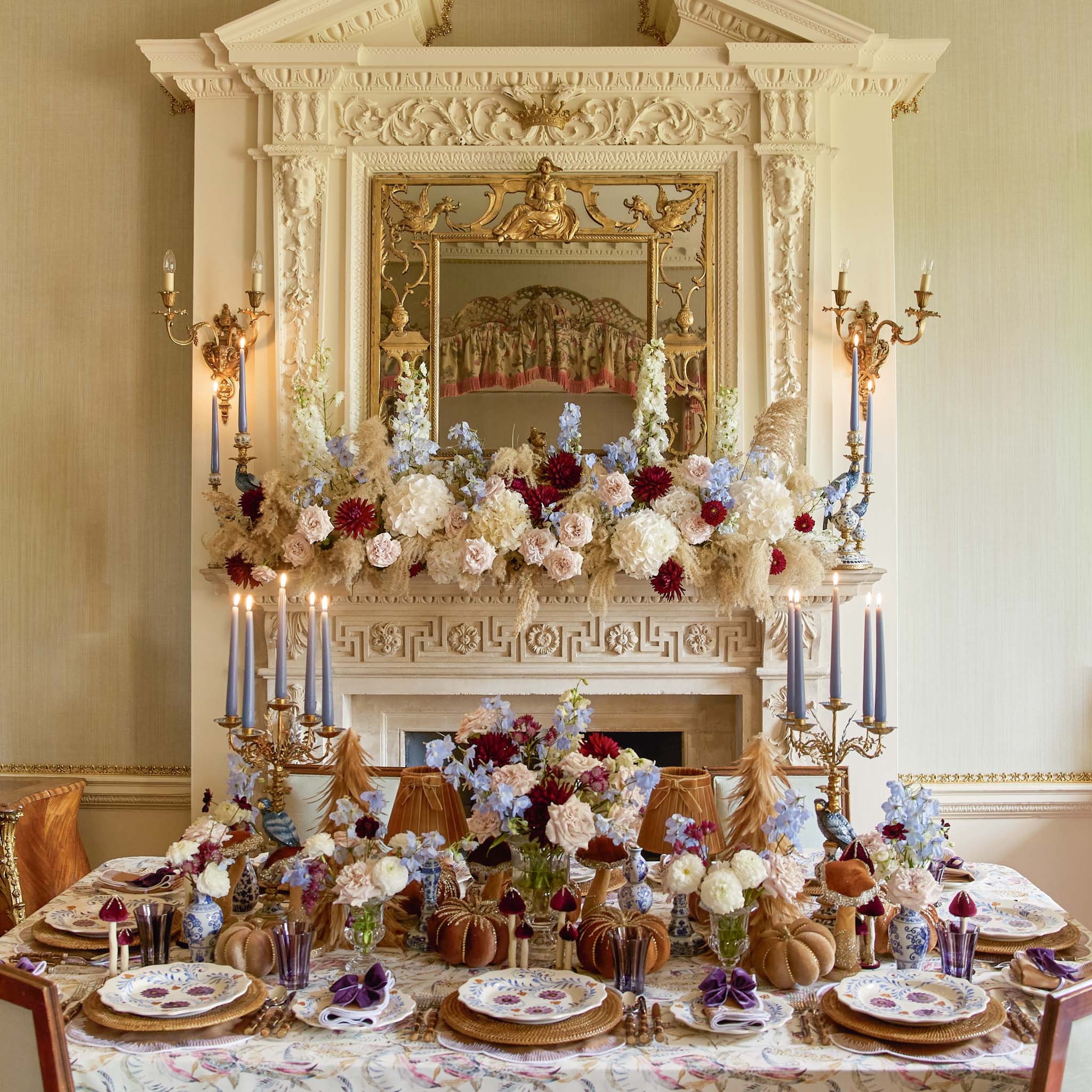 Decorative table setting with floral arrangements (Chinoiserie Pumpkin) and candles in a room with ornate fireplace and mirror.