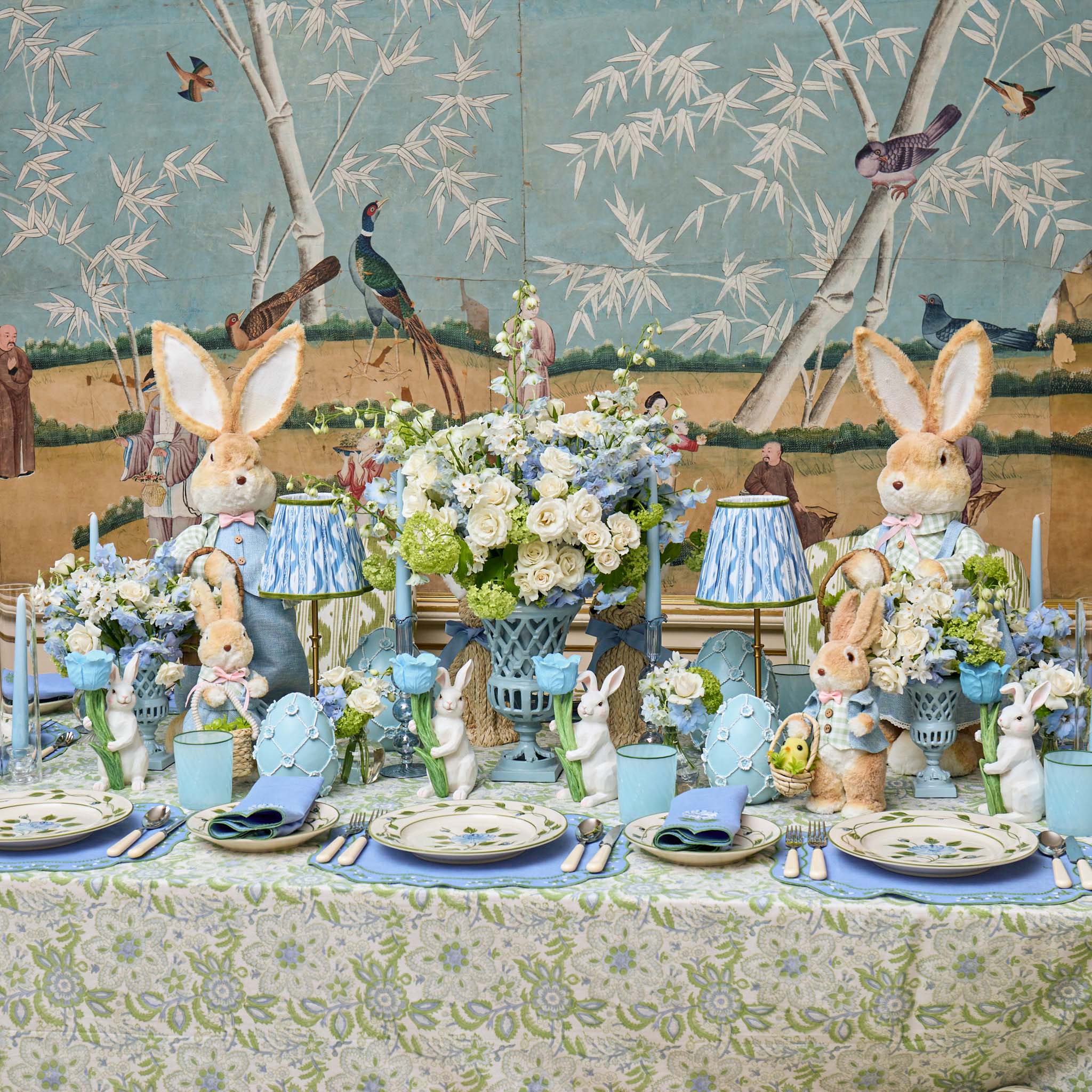 Easter-themed table setting with decorative rabbits and floral arrangements against a nature-themed wallpaper.