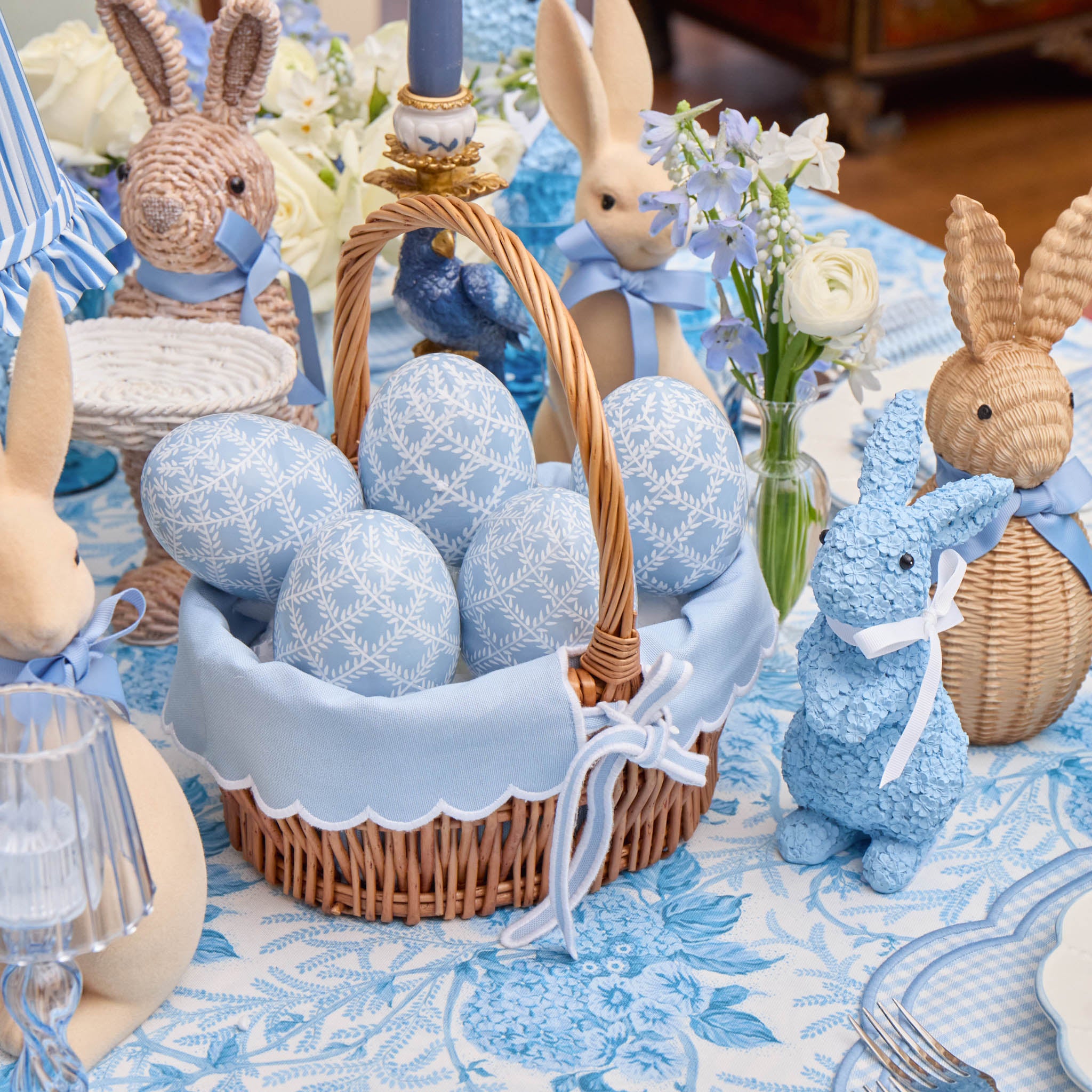 Decorative Easter scene with wicker basket of blue eggs, bunny figurines, and floral arrangement on a tablecloth.