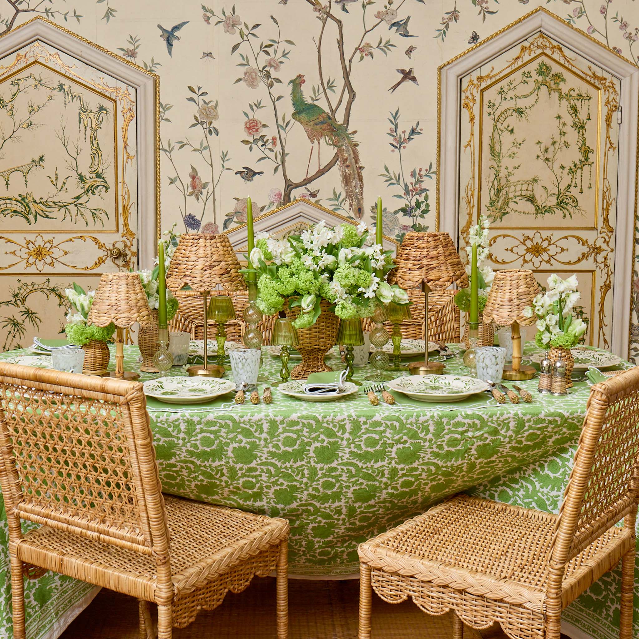 Dining table set with a green and white patterned tablecloth, wicker chairs, and decorative elements in a room with ornate wallpaper.
