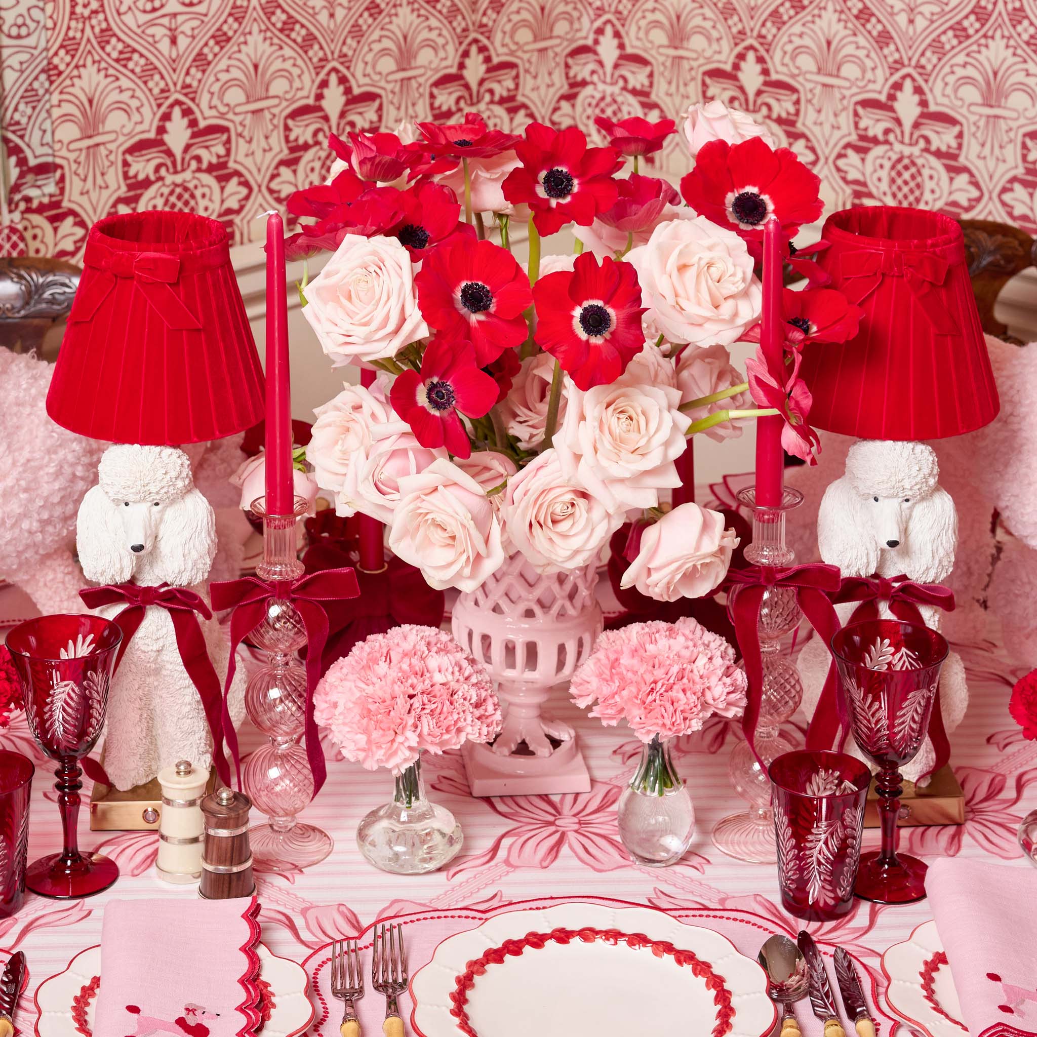 Decorative table setting with red and white flowers, candles, and small white dogs on a patterned background.