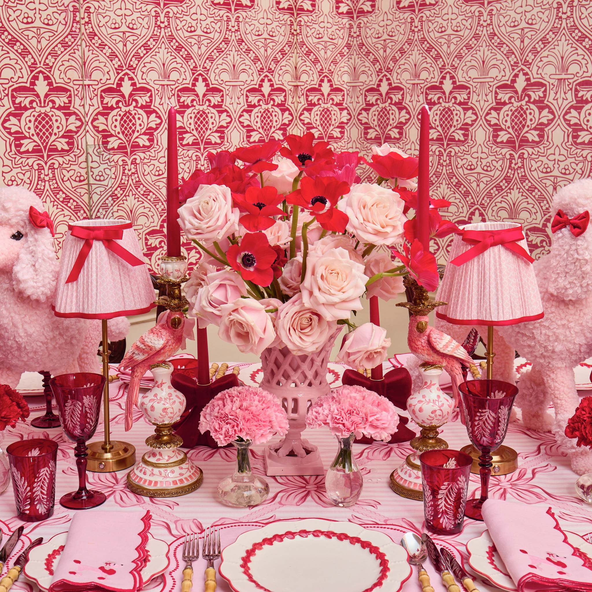 Decorative table setting with pink and red flowers, candles, and tableware against a patterned wall.