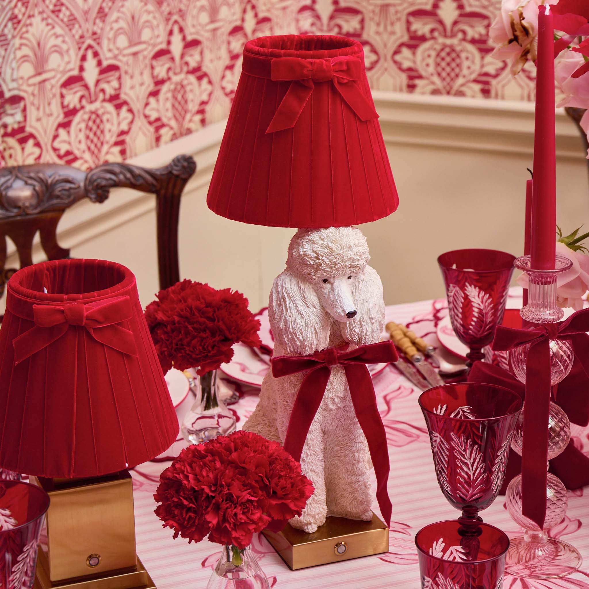 Decorative table setting with red lampshades and a poodle-shaped lamp on a patterned wall background.