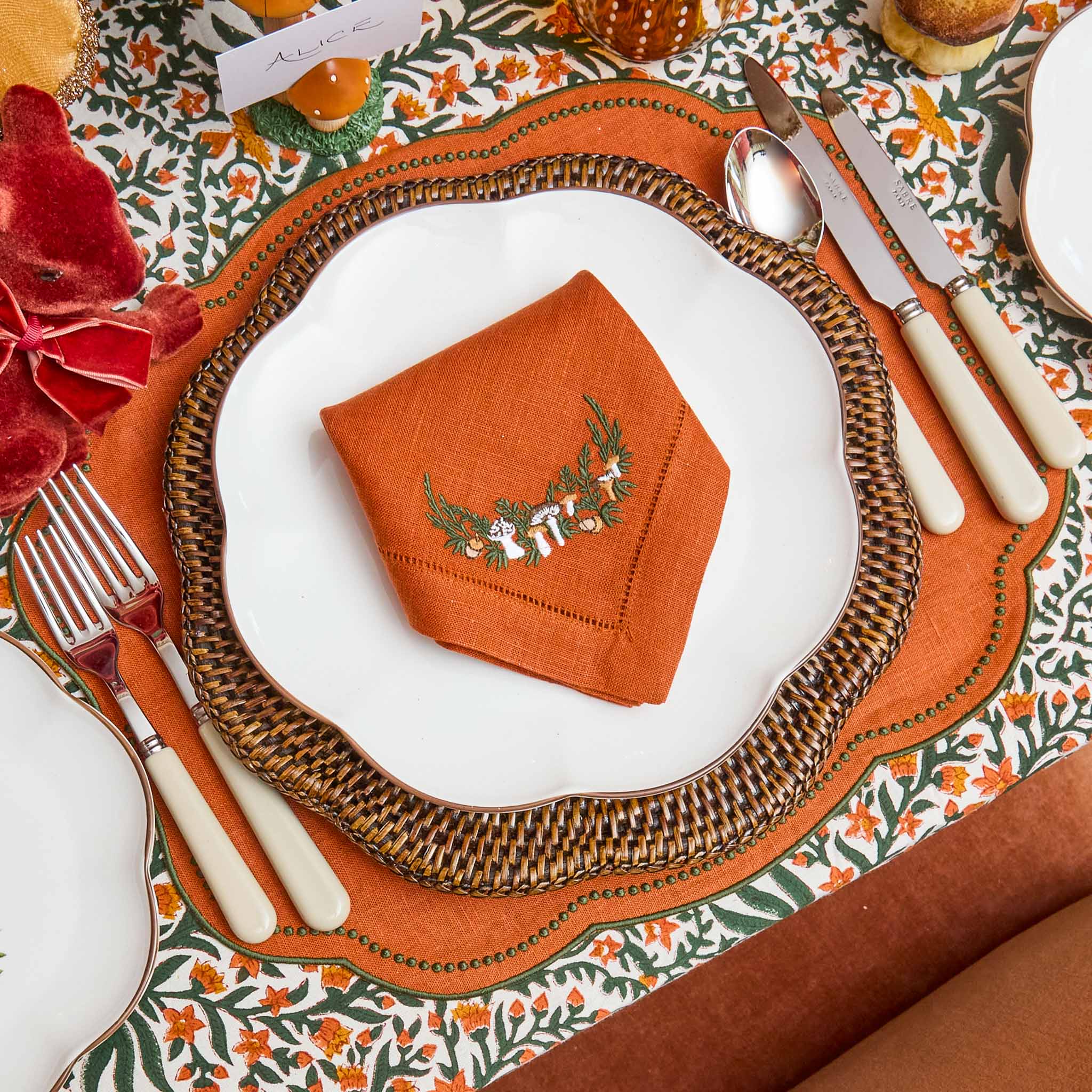 Decorative table setting with a mushroom embroidered orange napkin on a white plate, surrounded by patterned tablecloth and cream coloured cutlery.