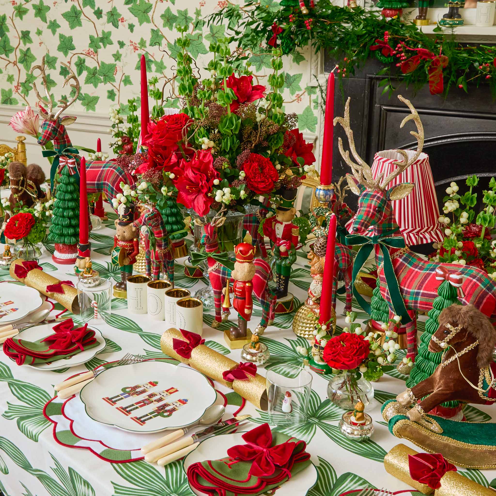 Decorative Christmas table setting with red and green ornaments, candles, and a fireplace in the background.