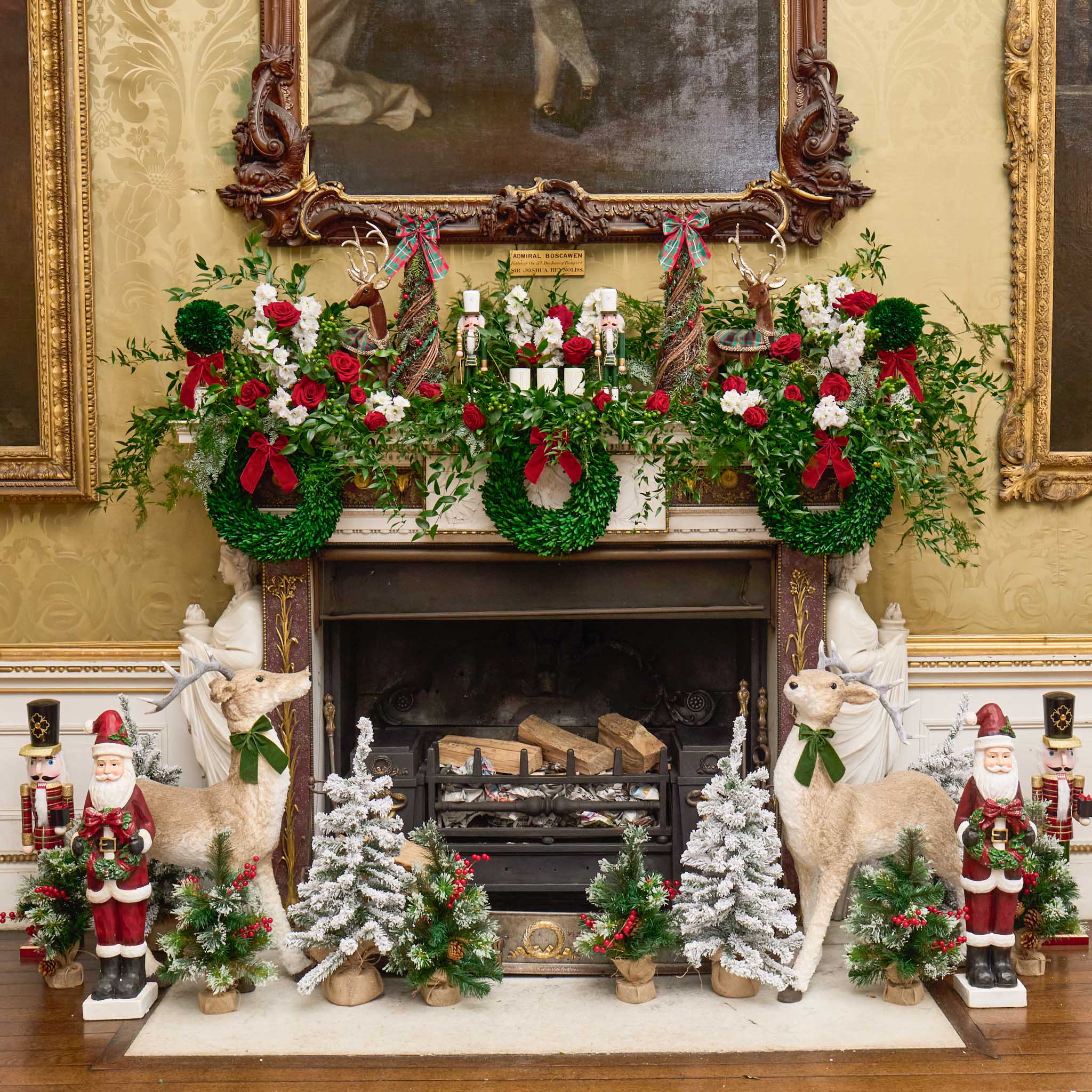 Decorative Christmas mantel with wreaths, trees, and figurines in a formal setting.