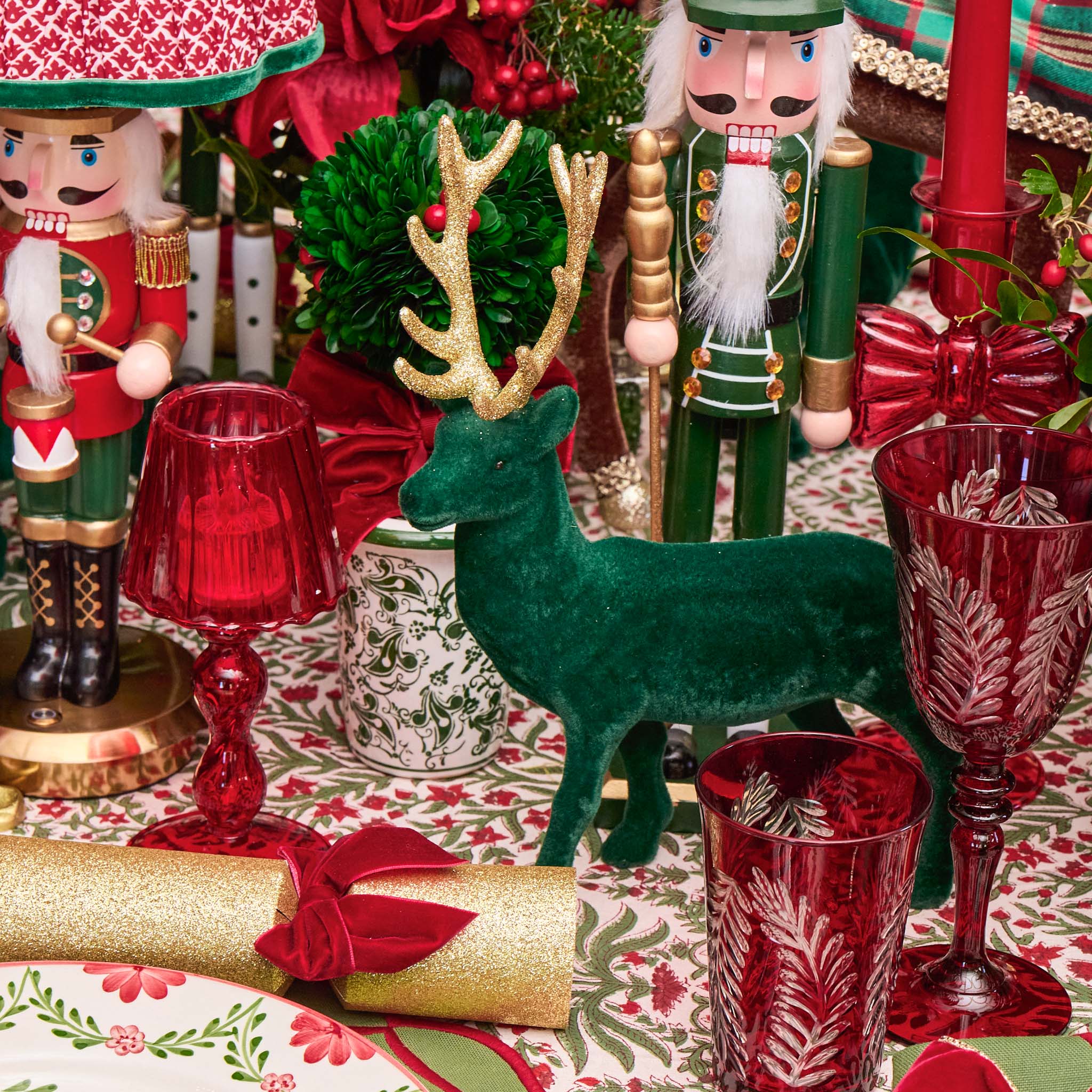 Decorative Christmas scene with nutcrackers, reindeer, and red glassware on a patterned tablecloth.