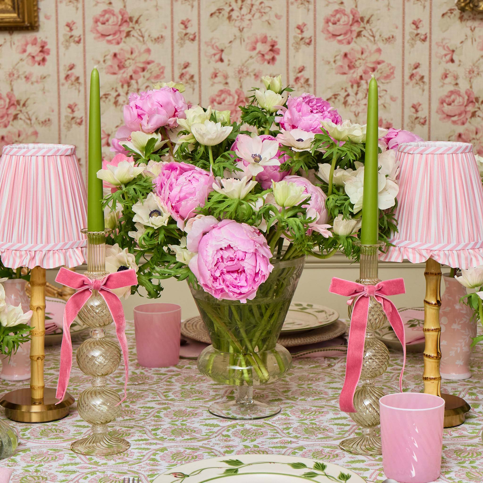 Decorative table setting with pink and green flowers, candles, and lamps against a floral wallpaper background.