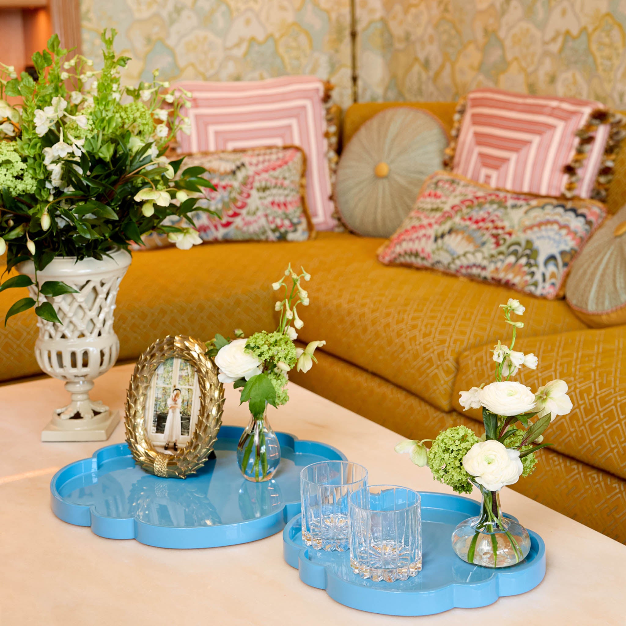 Decorative setting with flowers, mirror, and glasses on a blue tray in front of a yellow sofa with patterned cushions.