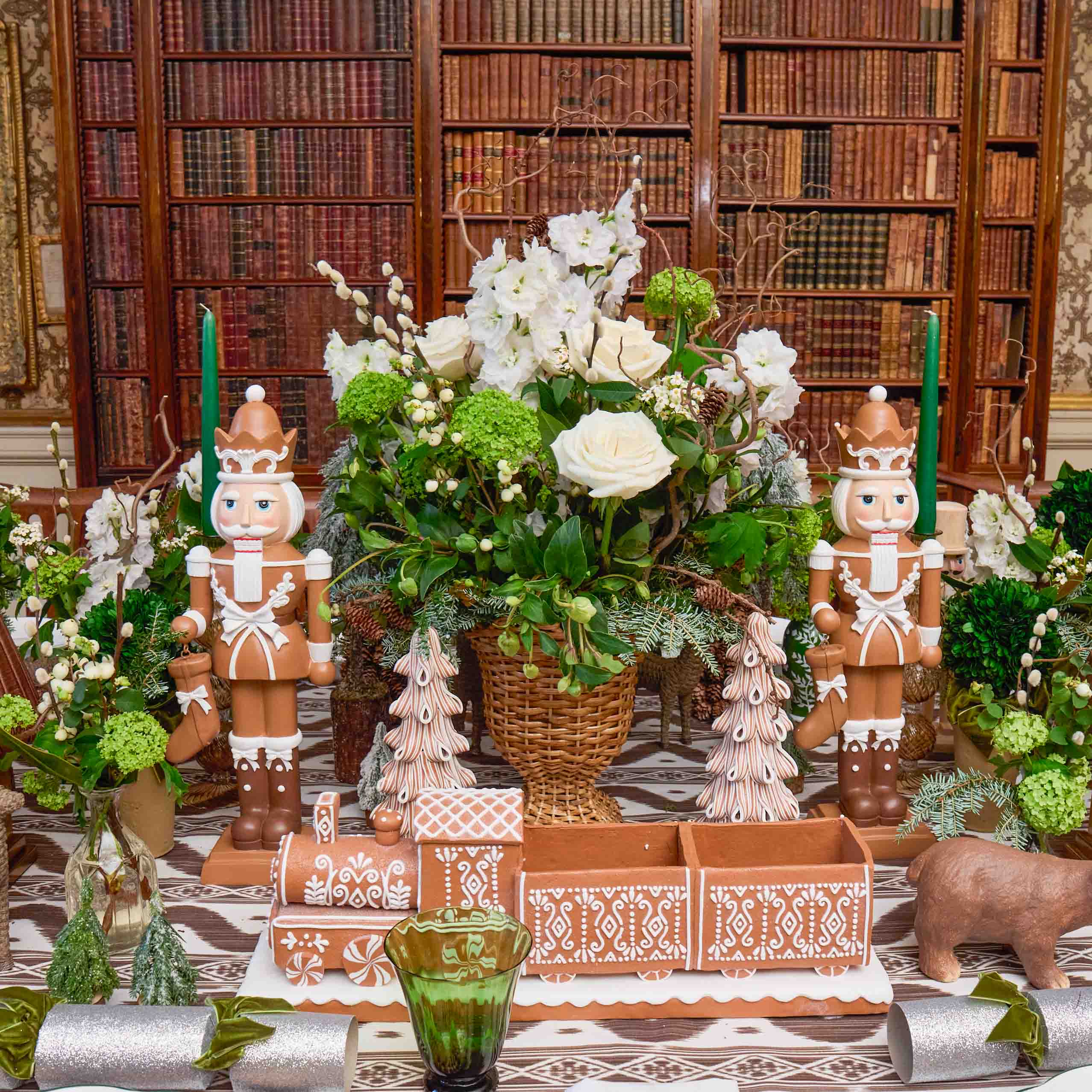 Decorative table setting with nutcrackers, floral arrangement, and gingerbread men in a room with bookshelves.