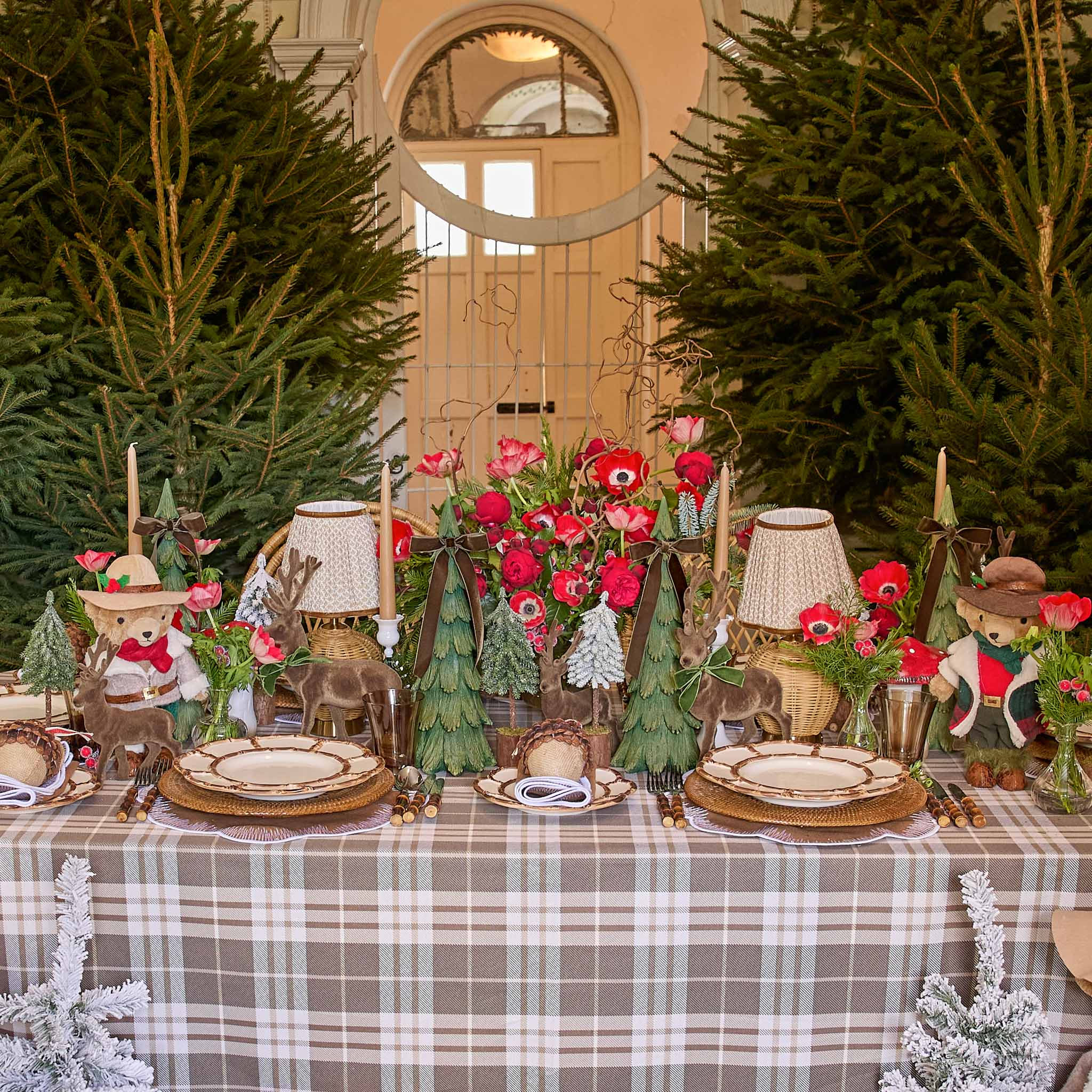 Decorative Christmas table setting with teddy bears, flowers, and small trees in a room with large windows.