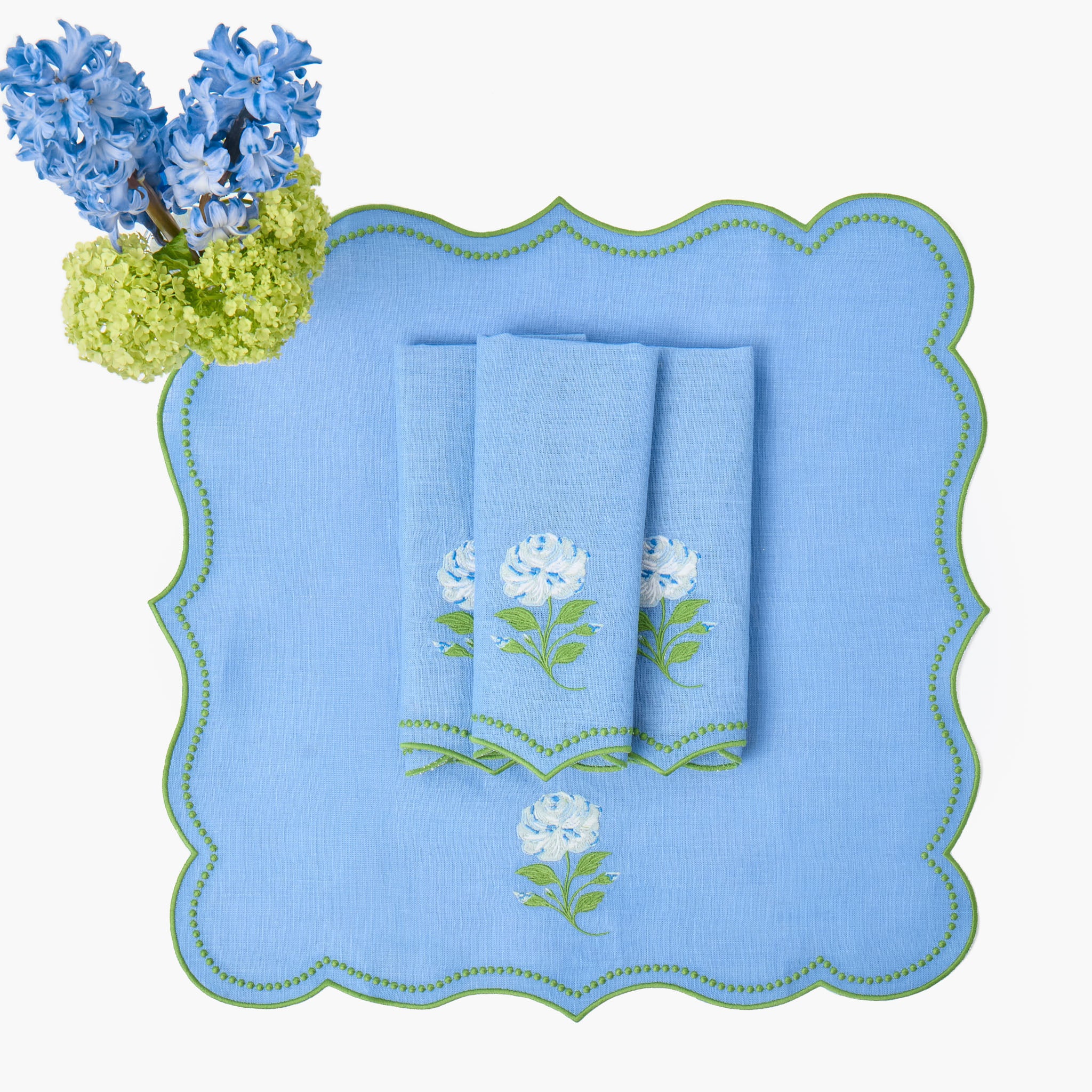 Set of blue napkins with floral embroidery on a matching placemat, accompanied by a bouquet of blue flowers.