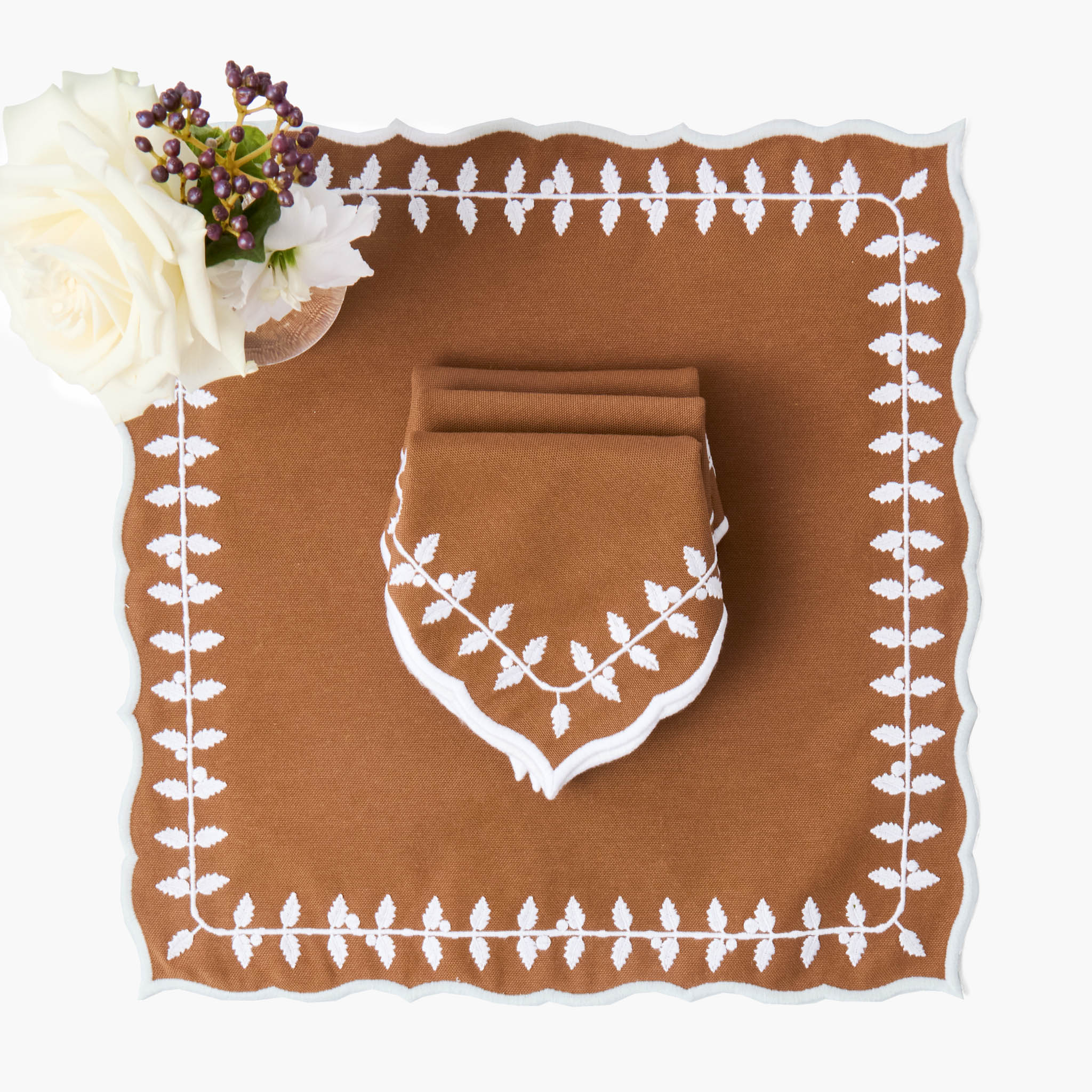 Brown placemat with white floral border and folded napkin, accompanied by a white rose.