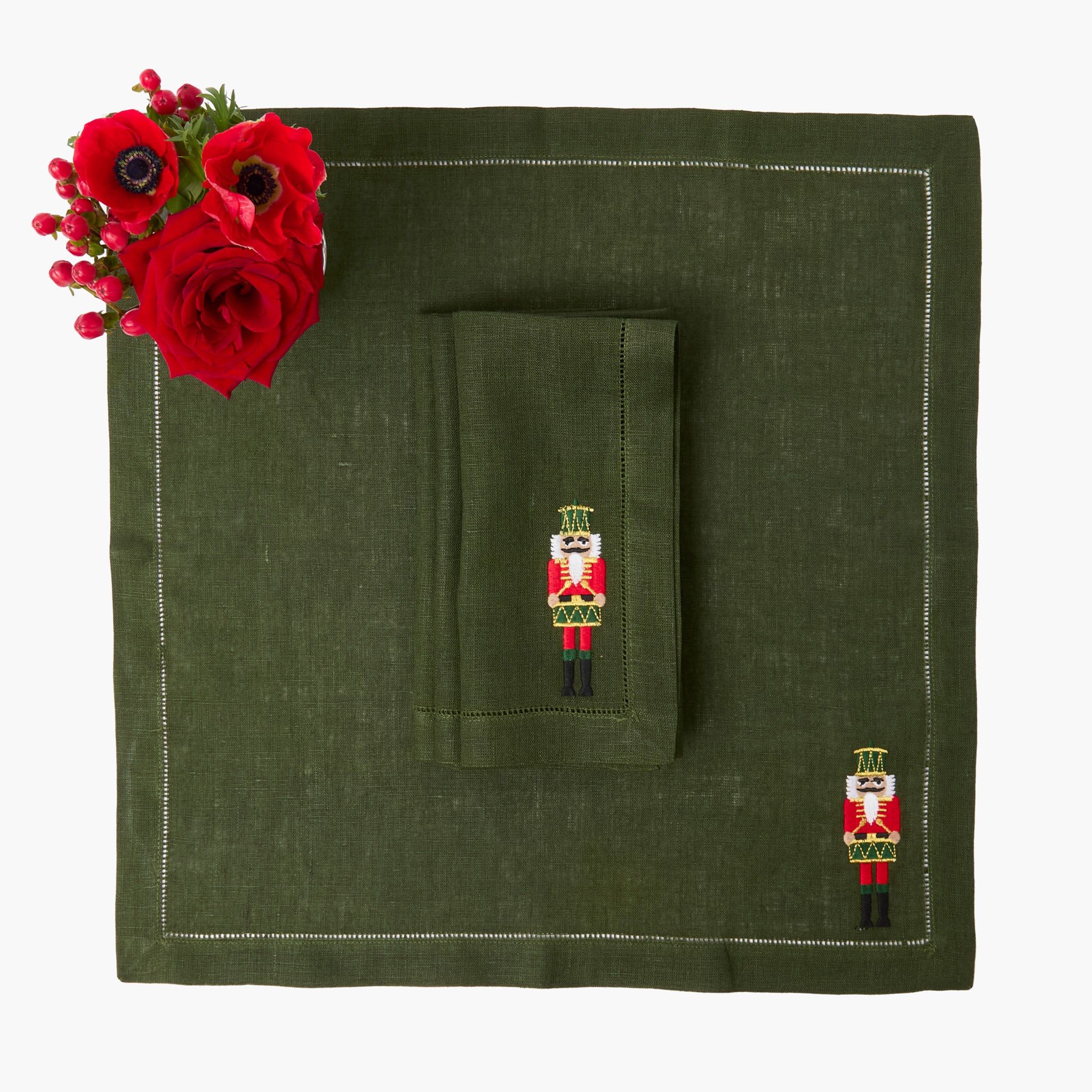 Green linen napkin with embroidered nutcracker design on a matching green placemat, accompanied by red flowers.